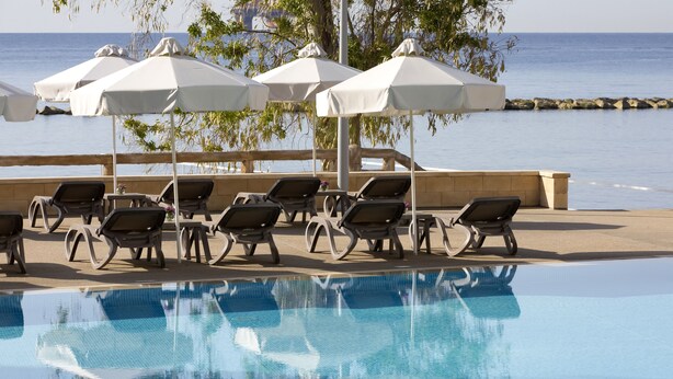 A poolside area with lounge chairs and umbrellas, a clear pool, and a view of the sea. - Harmony Bay Hotel (Limassol)