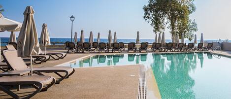 Zona de piscina con sillones de descanso y sombrillas, con vista despejada al mar y una piscina con baldosas.