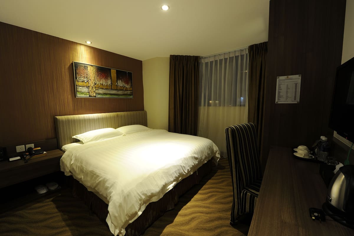 A hotel room with a large bed, a desk with a chair, a painting on the wall, and a window with curtains.