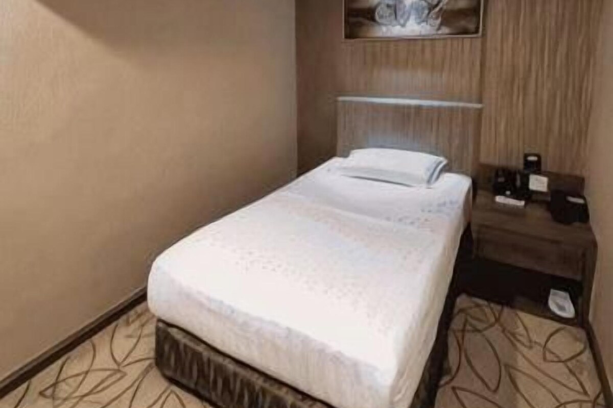 A hotel room with a bed, a bedside table, a wall-mounted air conditioner, and a decorative wall piece.