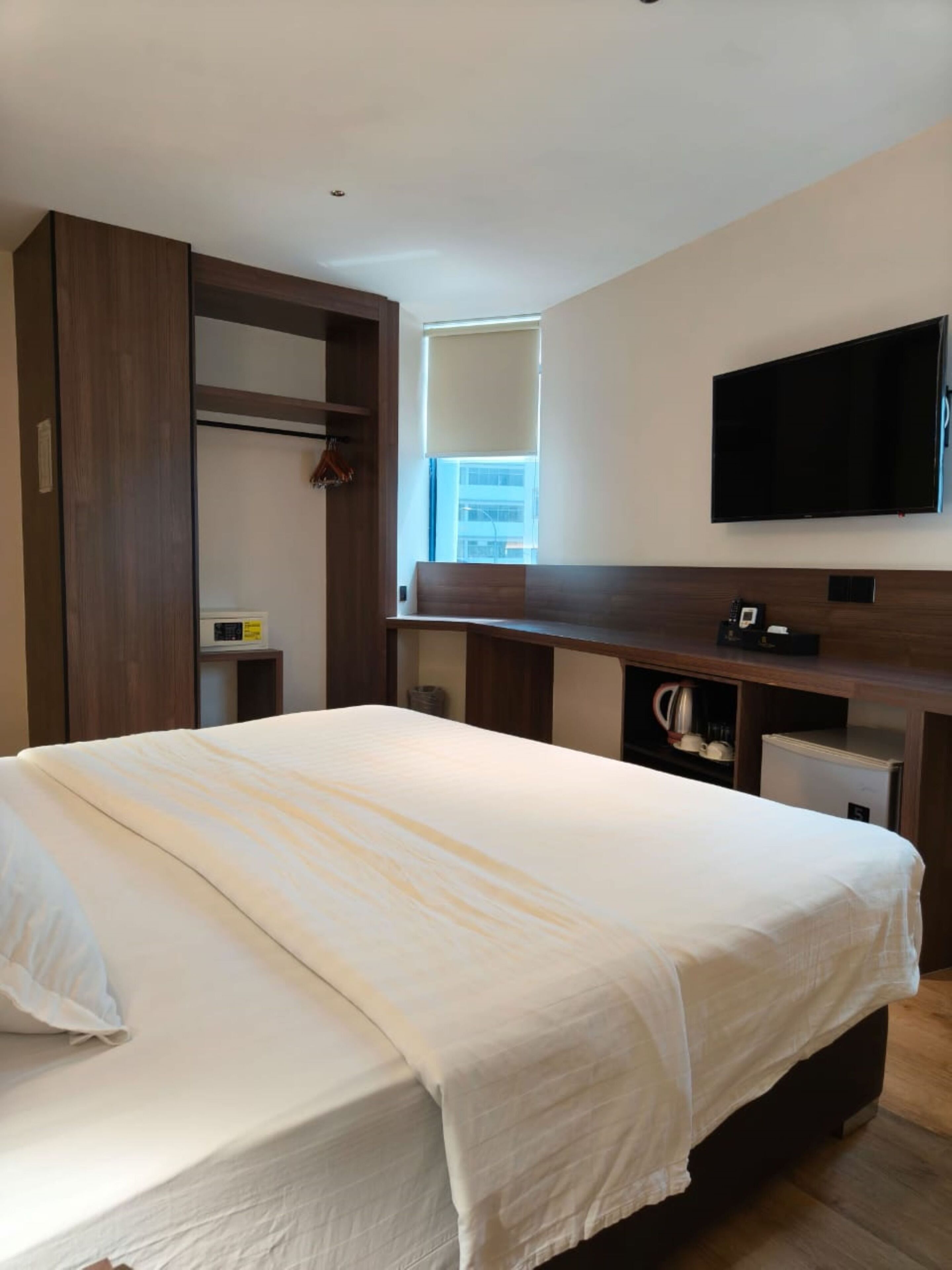A hotel room with a large bed, a TV mounted on the wall, a window with blinds, and a wooden wardrobe.
