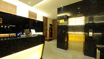 A hotel reception area with a modern design, featuring a reception desk, an elevator, and a luggage cart.