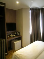A hotel room with a bed, a TV on a wooden shelf, a small fridge, and a window with curtains.