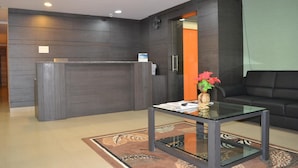 A reception area with a dark wood reception desk, a glass-top table with a vase of flowers, and a black leather sofa. - Shoba Inn (Bengaluru)