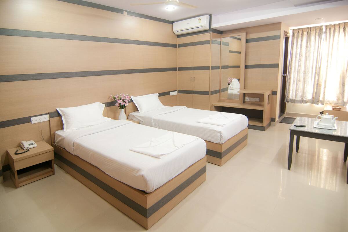 A hotel room with two beds, a bedside table with a lamp, a mirror, and an air conditioning unit.