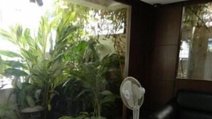 A room with a black leather sofa, a standing fan, and a large window with a view of greenery outside. - Shoba Suites (Bengaluru)