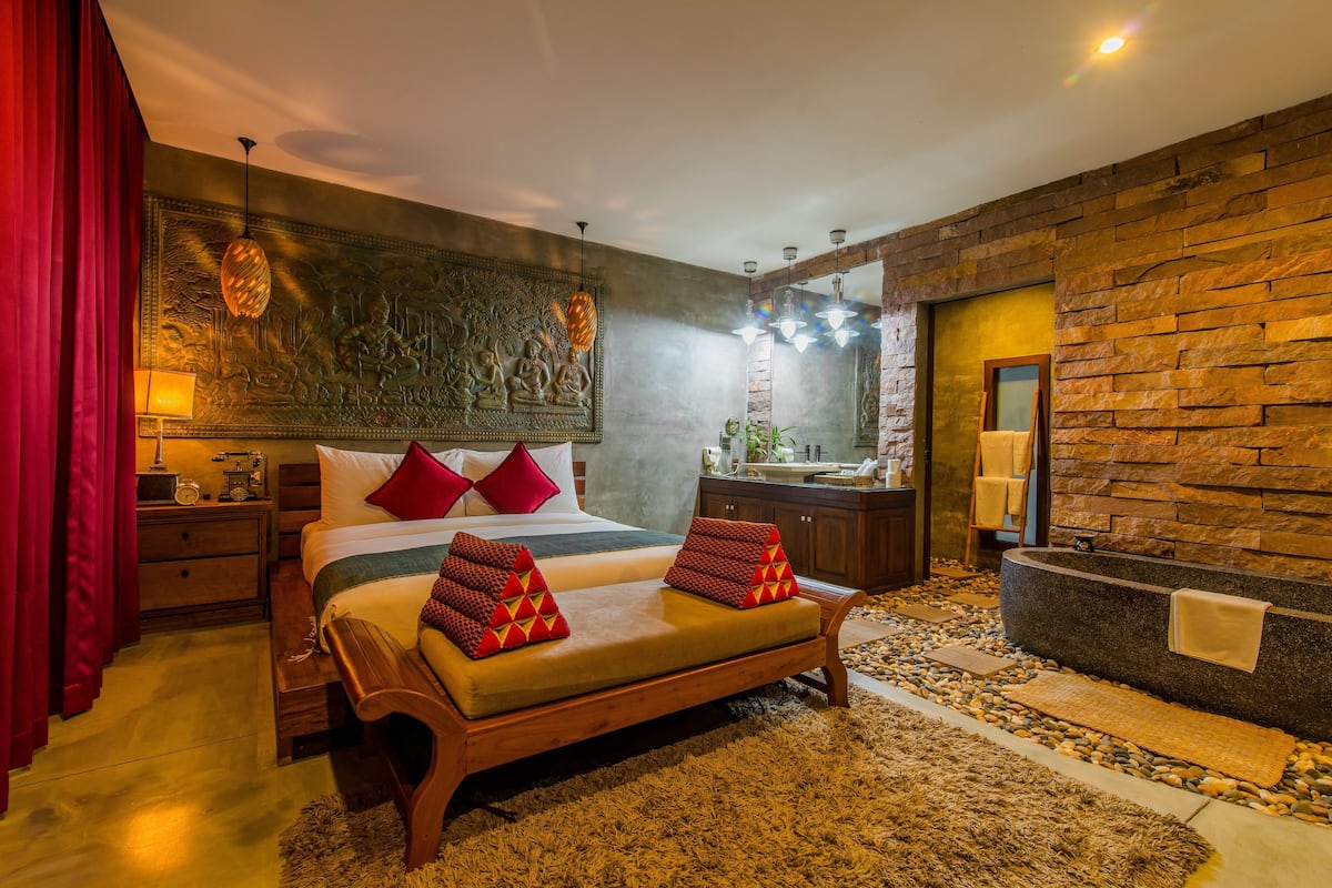 A modern hotel room with a large bed, a wooden bench, a stone wall, a bathtub, and a red curtain.
