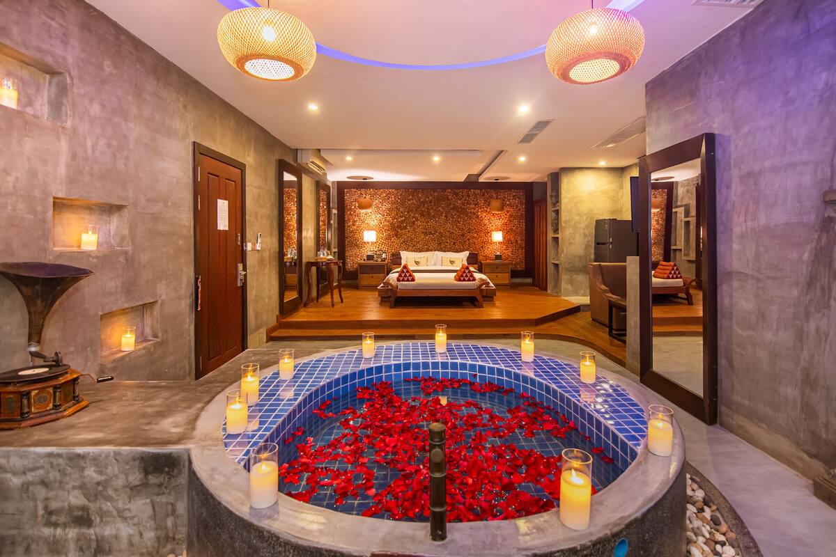 A luxurious spa room with a large bathtub filled with rose petals, a wooden bench, and a mirror reflecting the room.