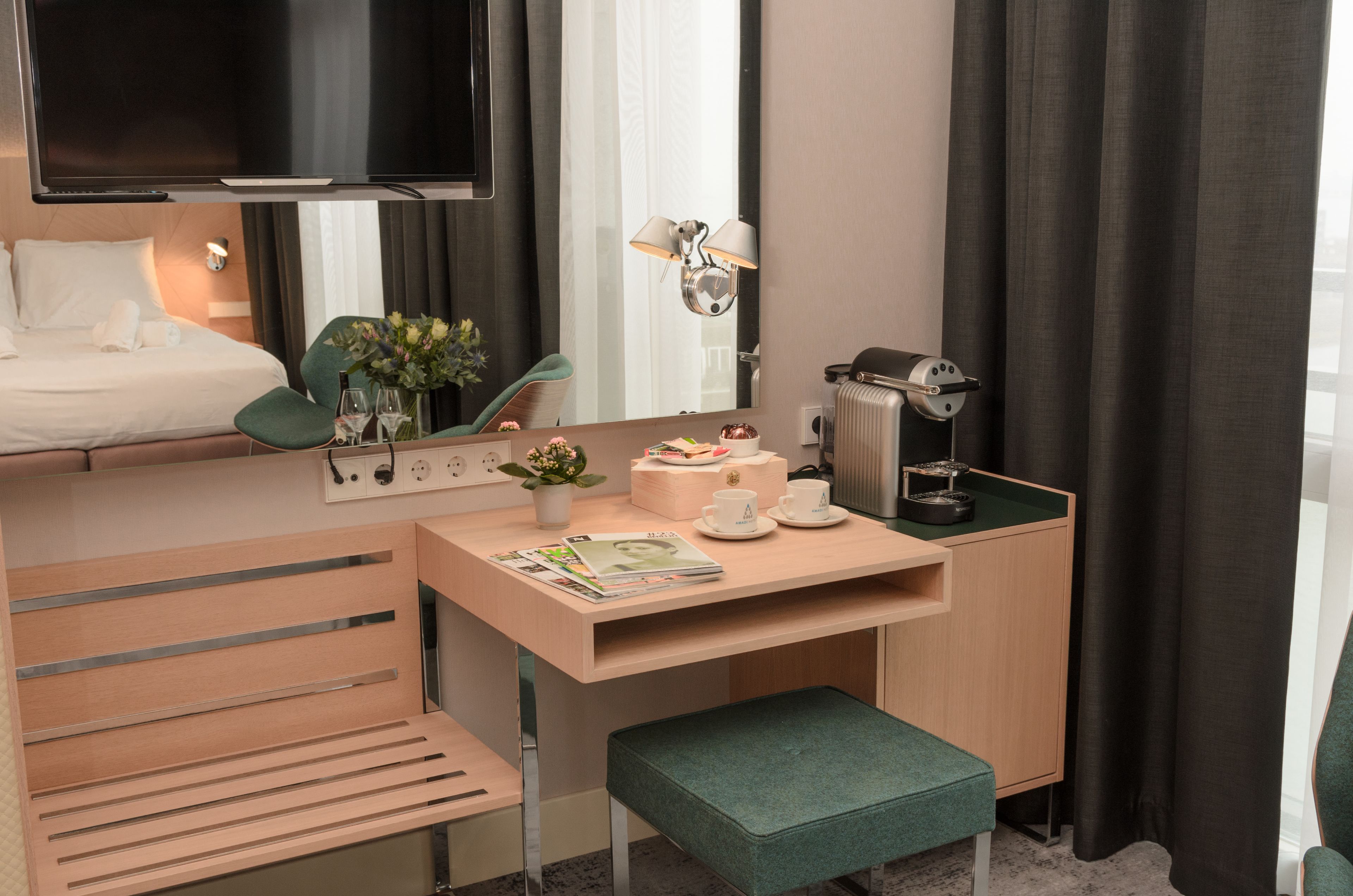 Ein gemütliches Hotelzimmer mit Bett, Schreibtisch mit Computer, Stuhl, kleinem Tisch mit Pflanze, Lampe und Heizkörper.
