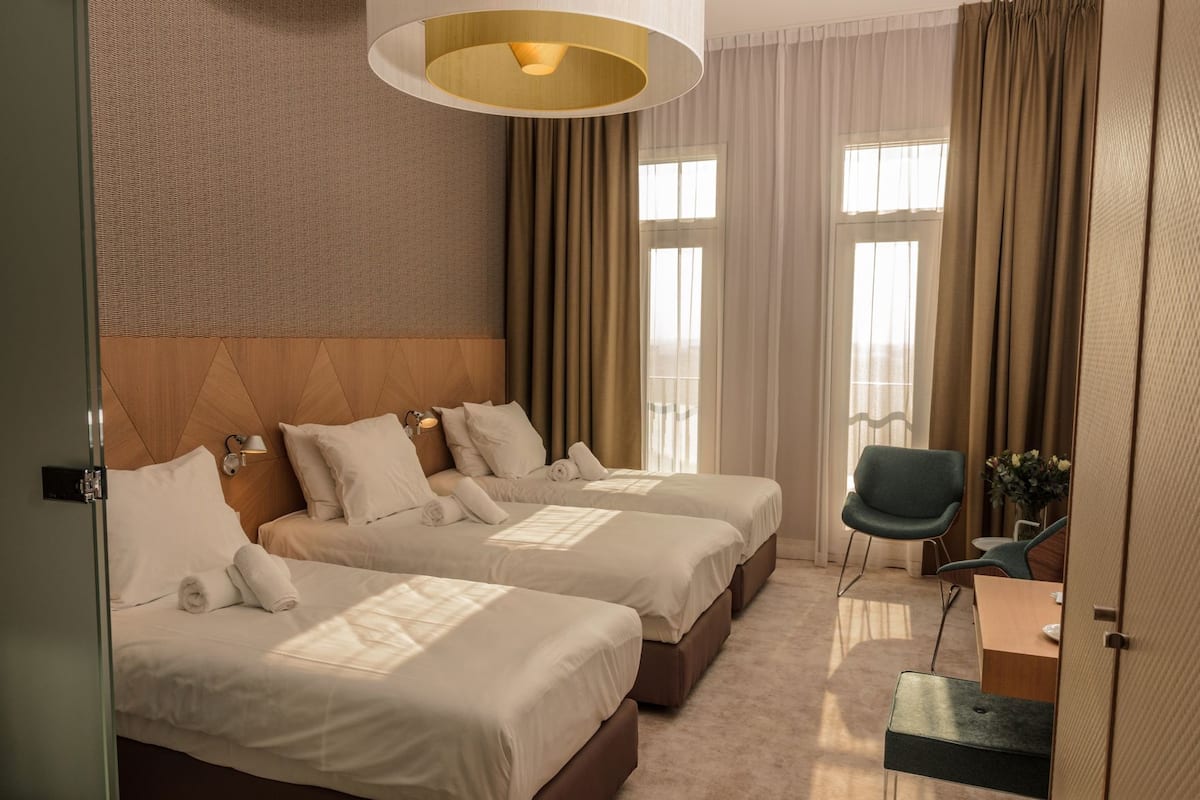 Ett hotellrum med två sängar, en sittgrupp med en fåtölj, ett skrivbord och ett stort fönster med gardiner.
