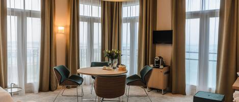 Habitación de hotel con una mesa de comedor redonda, cuatro sillas, un televisor de pantalla plana y un ventanal con cortinas.