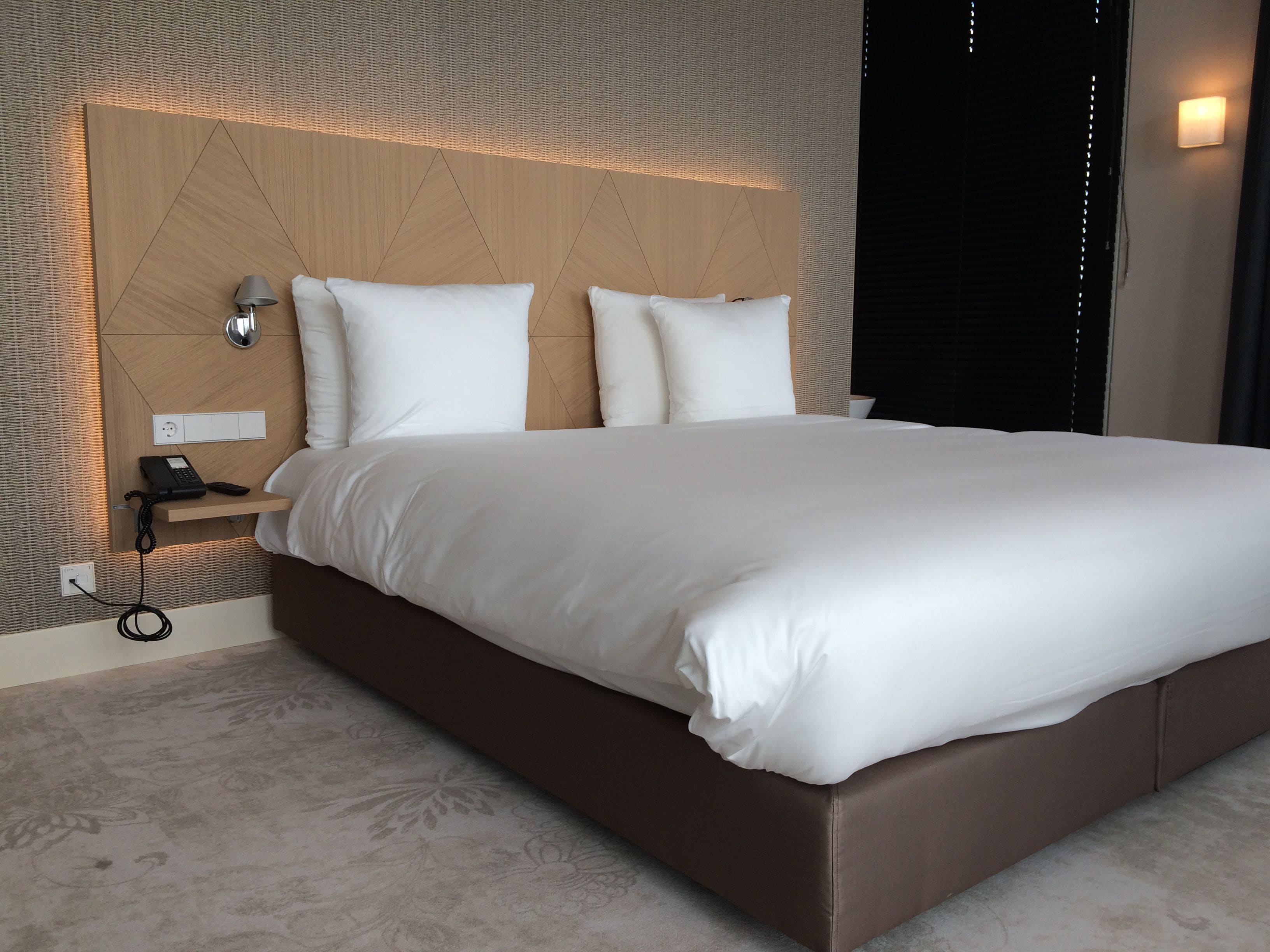 A modern hotel room with a large bed, a wooden headboard, a bedside table with a lamp, and a wall-mounted phone.