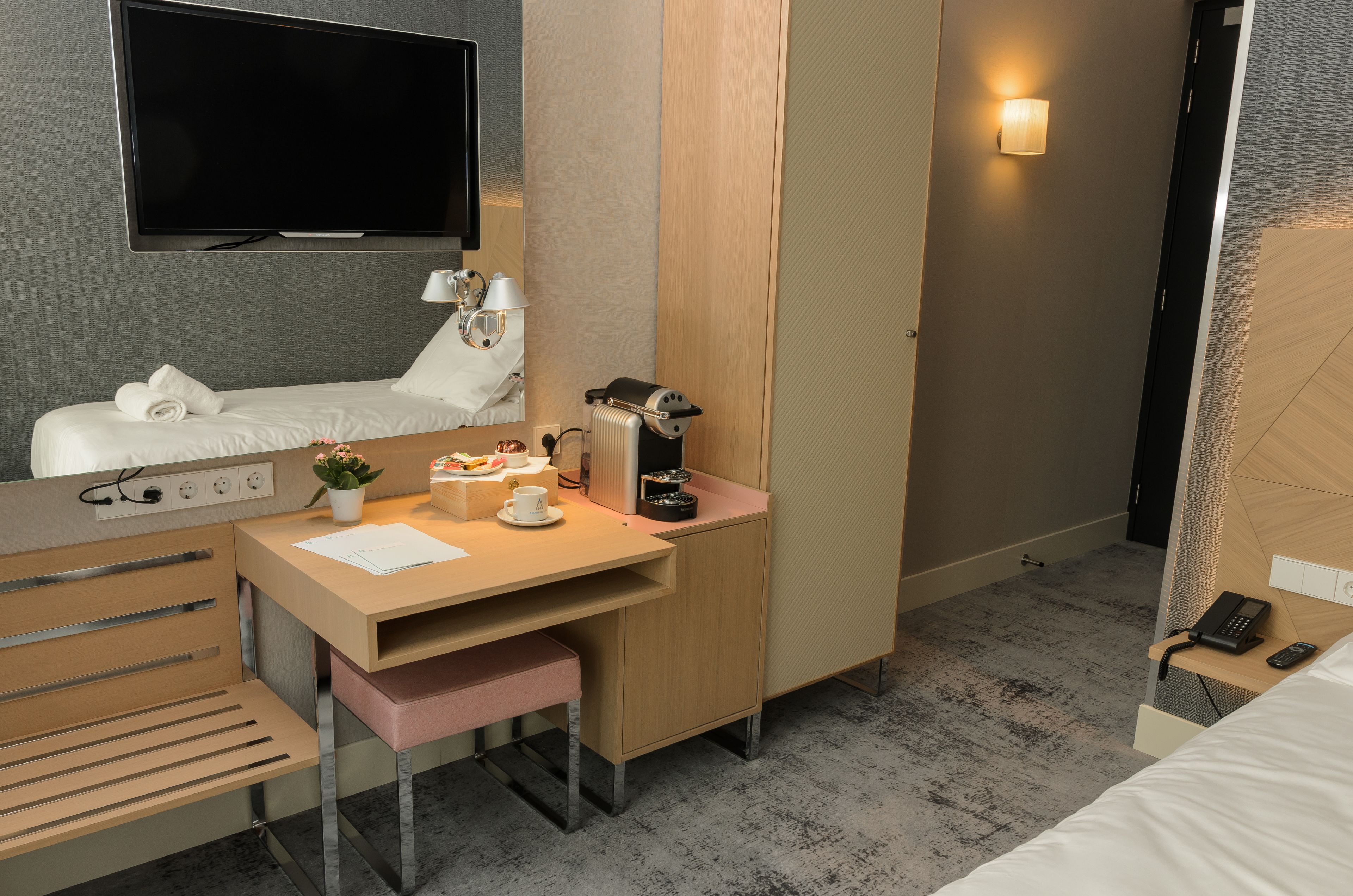 A hotel room with a bed, a desk with a computer, a TV, and a bedside table with a lamp and a phone.