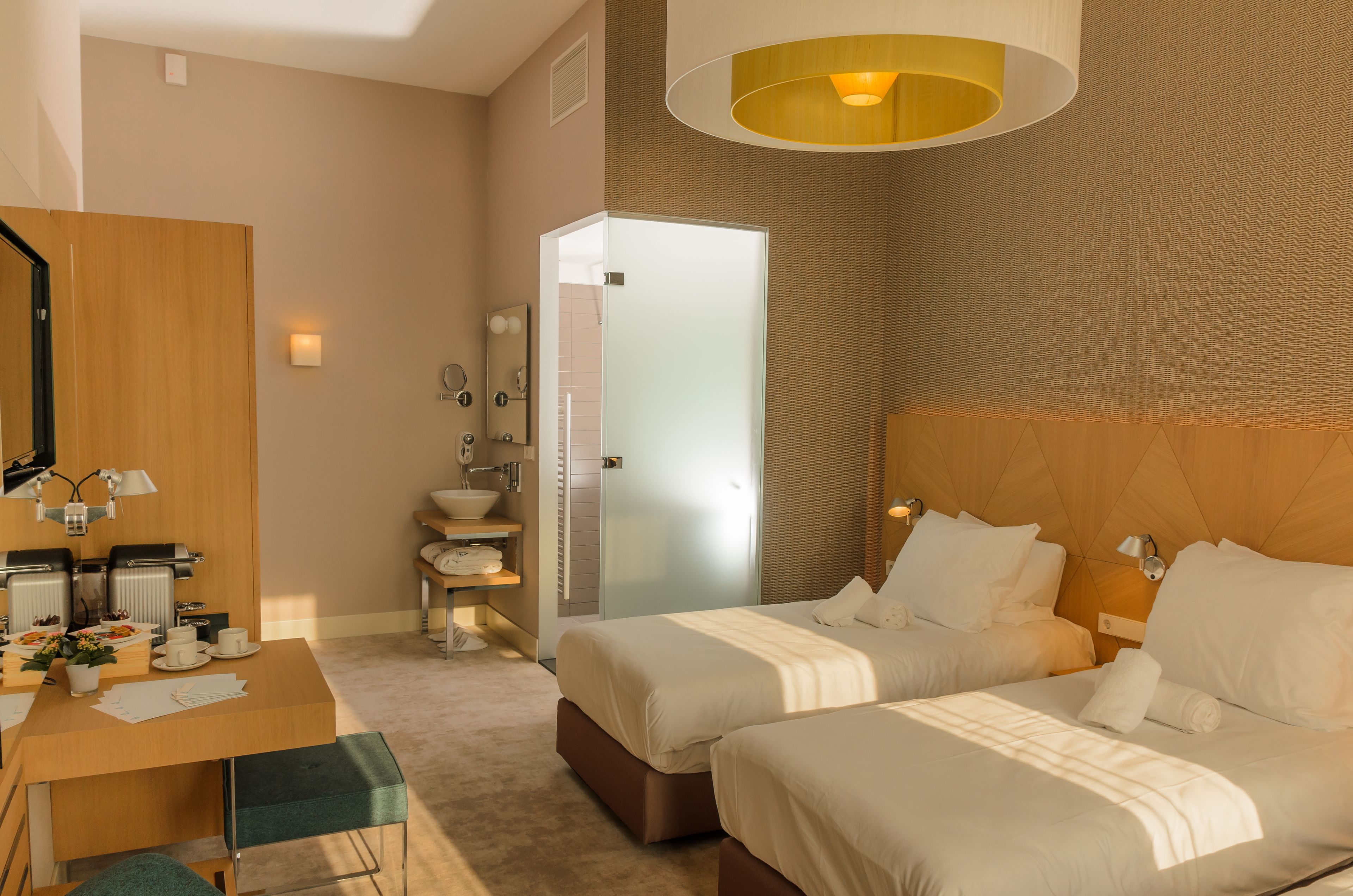 A hotel room with two beds, a wooden desk with a chair, a small bathroom, and a large window.