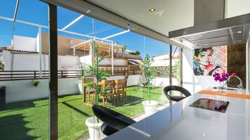 A modern kitchen with a large window, a dining area with a table and chairs, and a view of a garden and neighboring buildings.