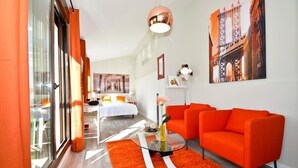 Une chambre d’hôtel moderne avec un grand lit, un coin salon avec des canapés orange, une table basse en verre et une grande fenêtre avec des rideaux.