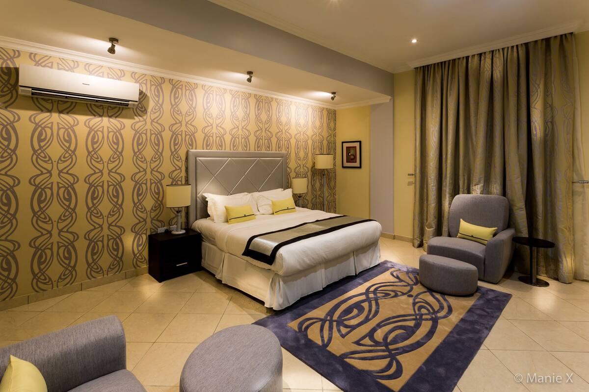 A modern hotel room with a large bed, a grey armchair, a small table, and a decorative rug.