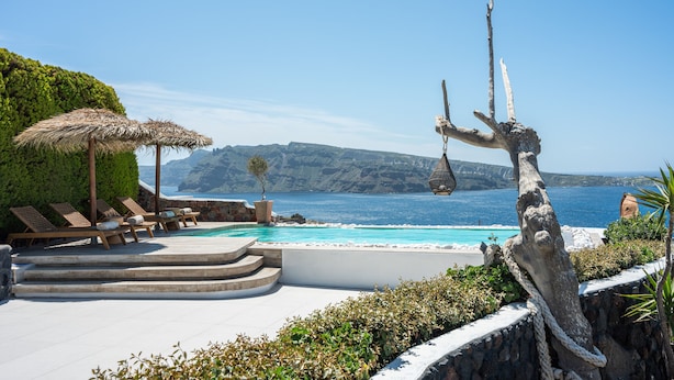 A poolside area with lounge chairs and a thatched umbrella, overlooking a clear blue sea and rocky cliffs.