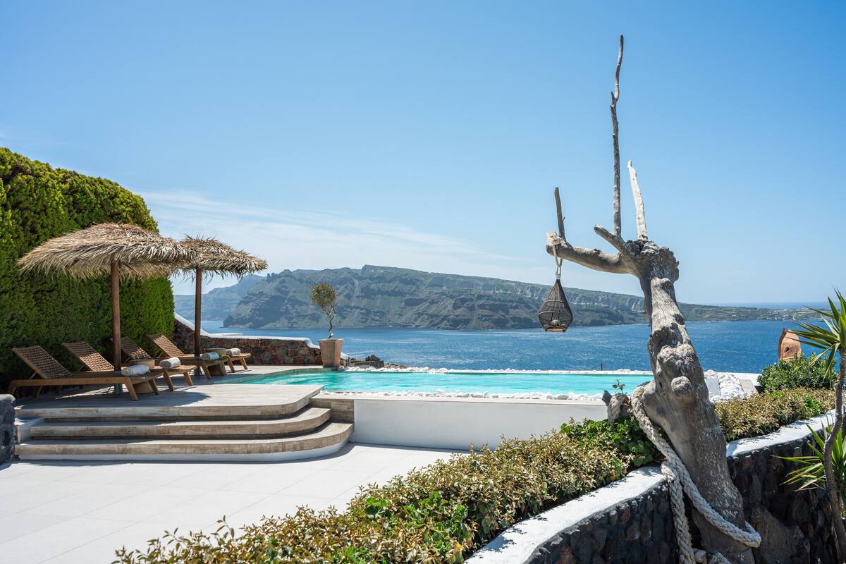 A poolside area with lounge chairs and a thatched umbrella, overlooking a clear blue sea and rocky cliffs.