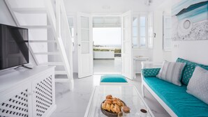 A modern living room with a television, a glass coffee table with pastries, a teal sofa, and a view of the beach through an open door.