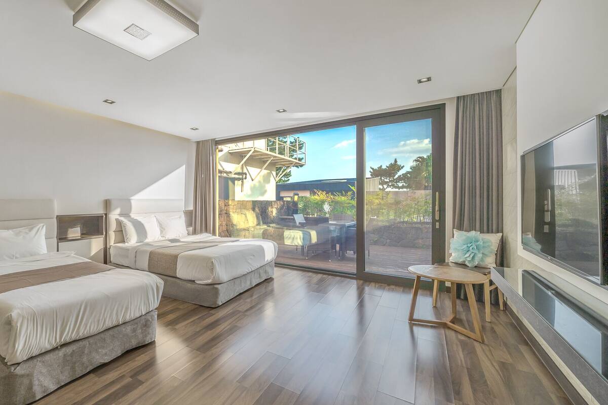 A modern hotel room with two beds, a large sliding glass door to a balcony, and a TV on the wall.