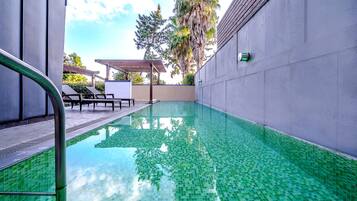 A swimming pool with a tiled floor, a pergola with chairs, and a wall-mounted light fixture.