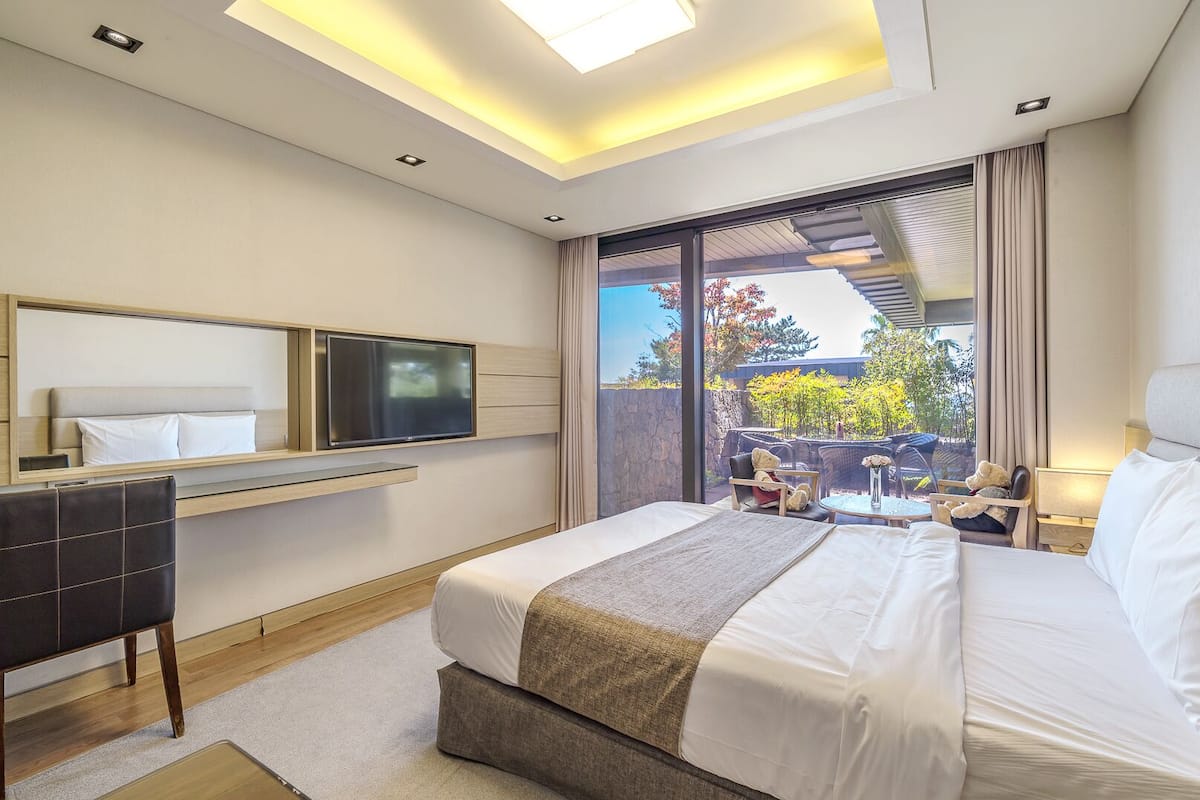 A modern hotel room with a large bed, a flat-screen TV mounted on the wall, a small seating area with a table and chairs, and a view of the outdoors through a large window.