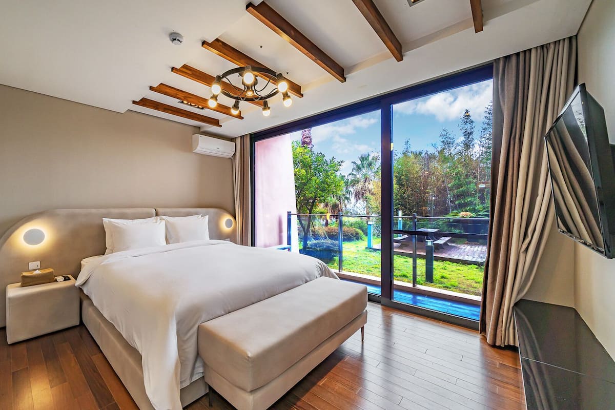 A modern bedroom with a large bed, a nightstand, a TV, and a balcony with a view of greenery.