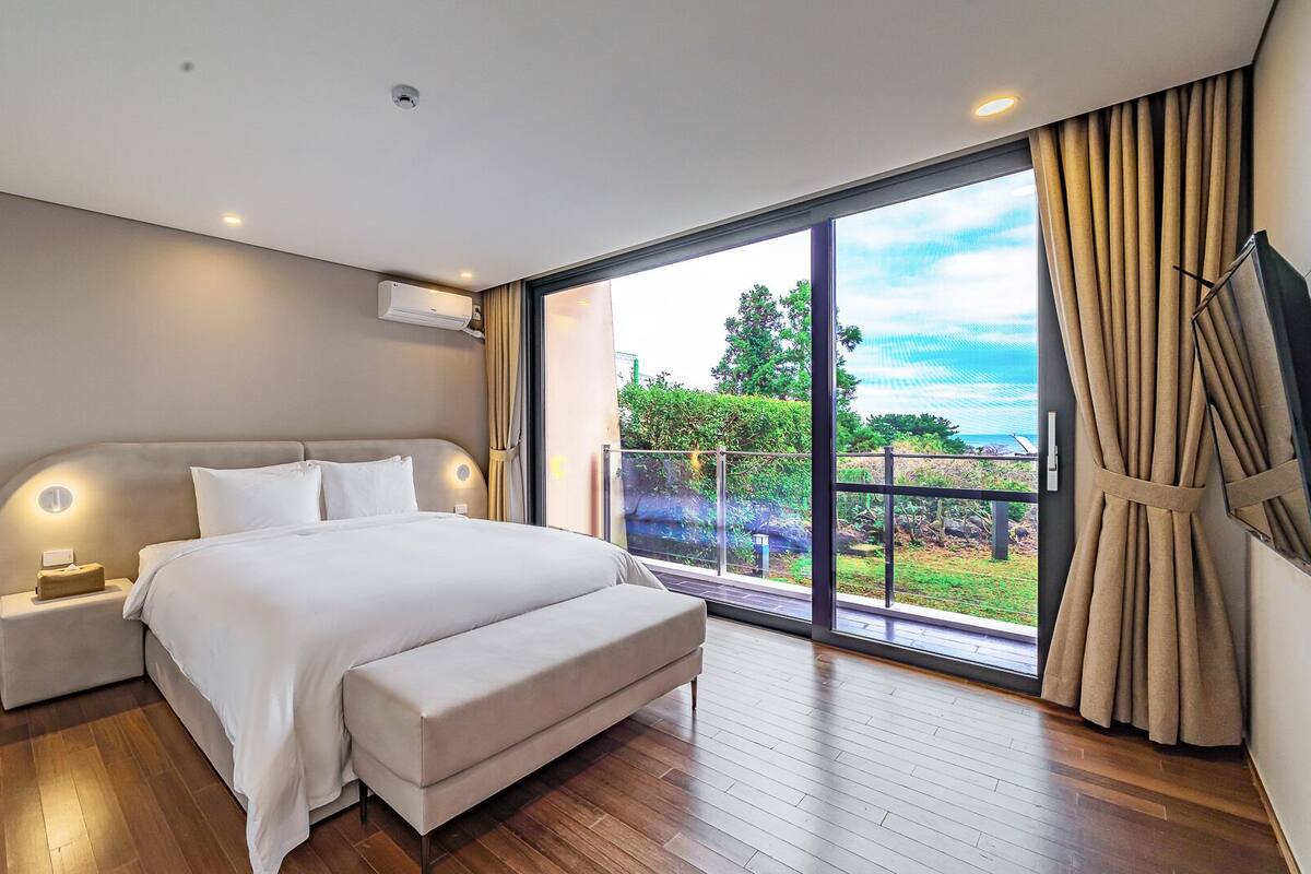 A modern bedroom with a large bed, a nightstand, a bench, and a sliding glass door leading to a balcony with a view of greenery and a body of water.