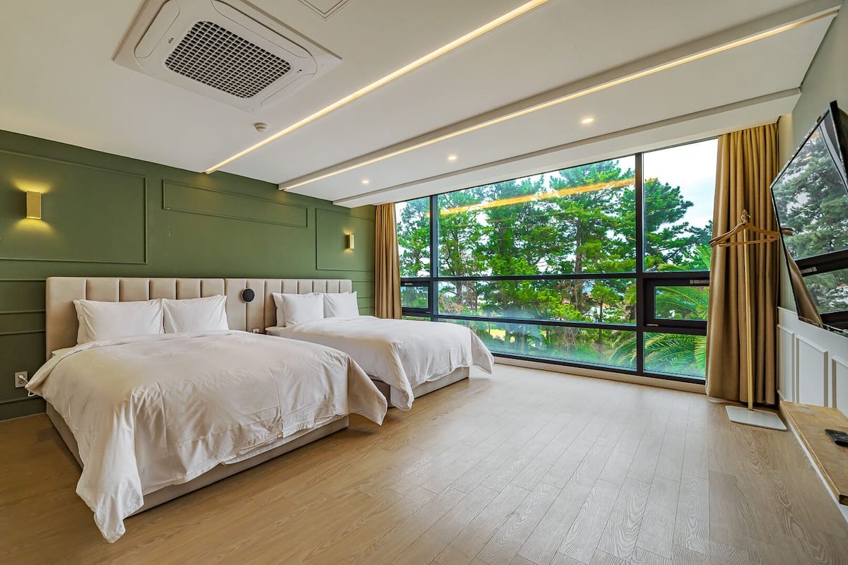 A modern bedroom with two beds, a large window with a view of trees, and recessed lighting.
