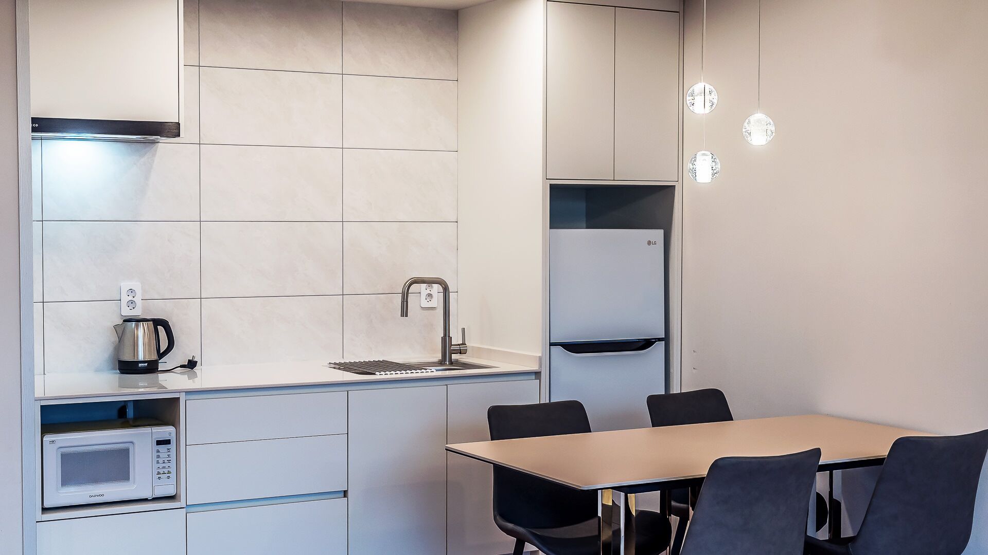 A modern kitchen with a microwave, kettle, and sink. There is a table with chairs and a cabinet with shelves.