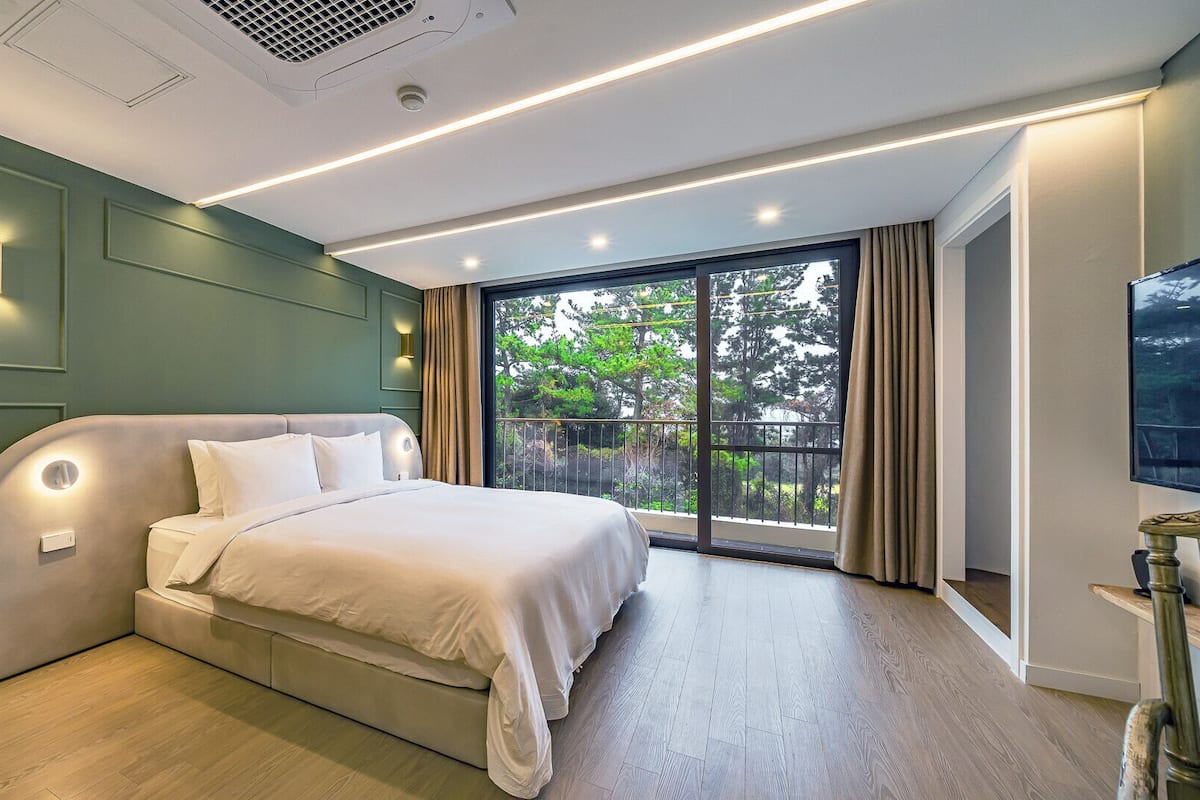 A modern bedroom with a large bed, built-in lighting, and a balcony with a view of trees.