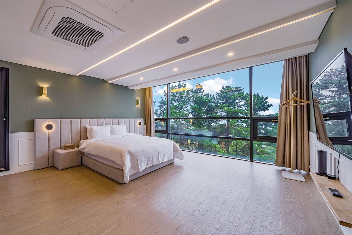A modern bedroom with a large window, a bed, a bedside table, and a TV mounted on the wall.