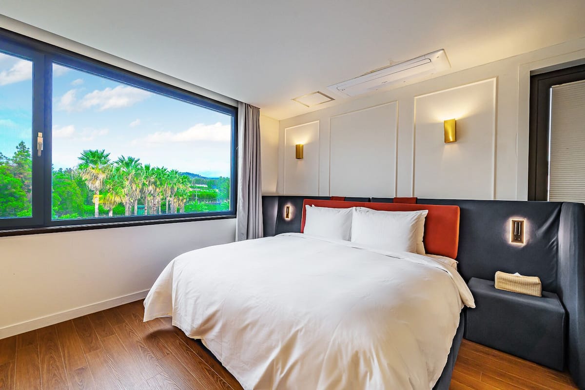 A hotel room with a large window, a bed with white bedding, a red and black headboard, and a wall-mounted light fixture.