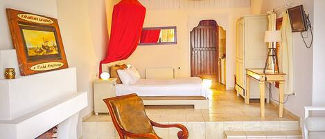A room with a bed, a wooden chair, a desk with a lamp, and a fireplace.