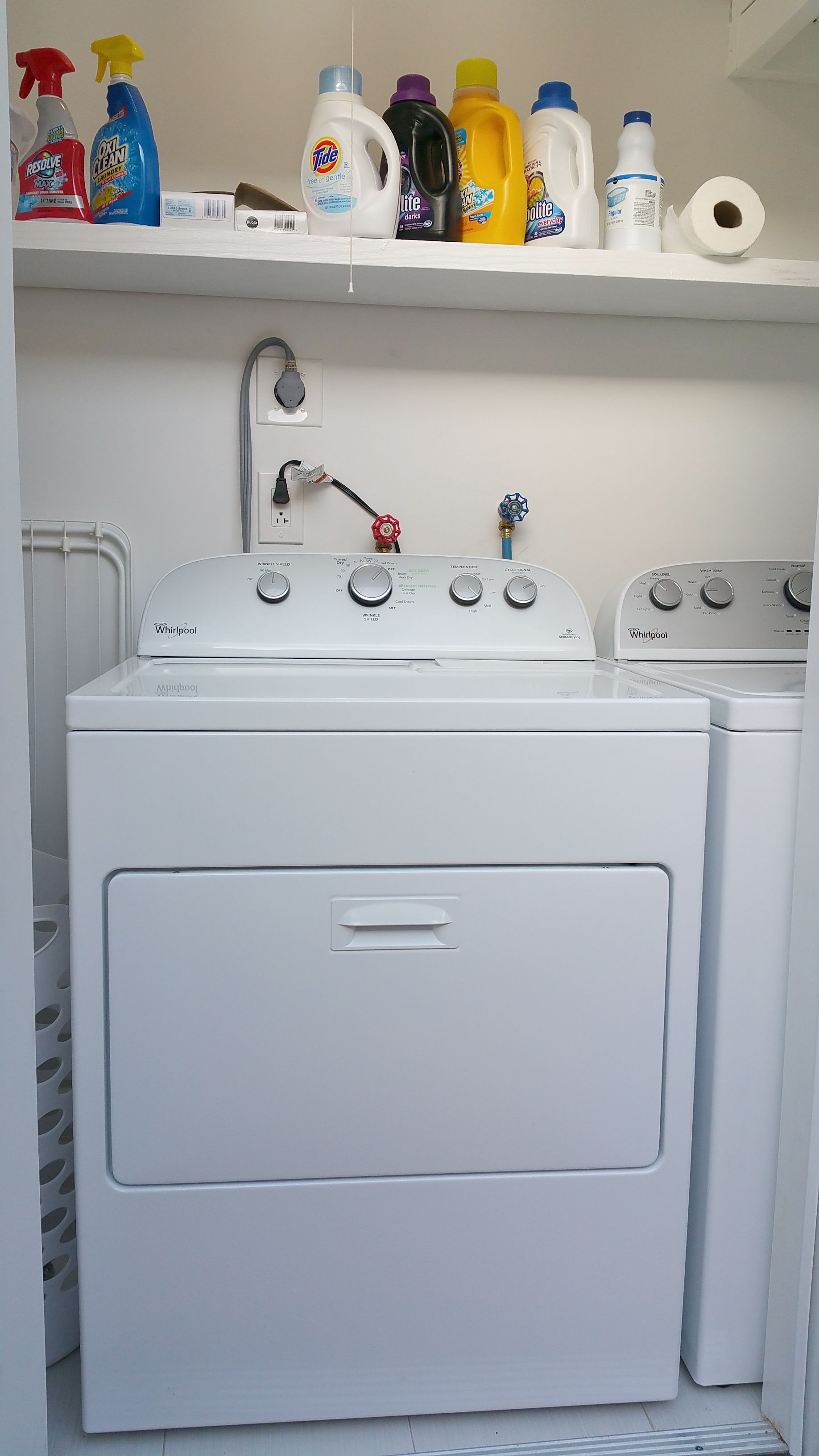 A laundry area with a Whirlpool washer and dryer, a shelf with various laundry detergents, and a roll of toilet paper.
