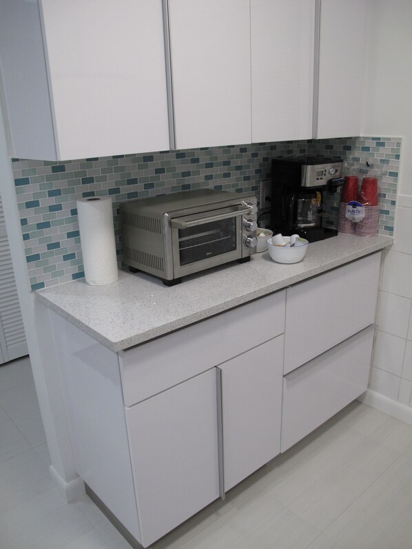Una cocina con gabinetes blancos, horno tostador, cafetera y vasos de plástico rojos.