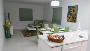 A modern kitchen with white cabinets, a dining area with green chairs, a white countertop, and a basket of snacks.