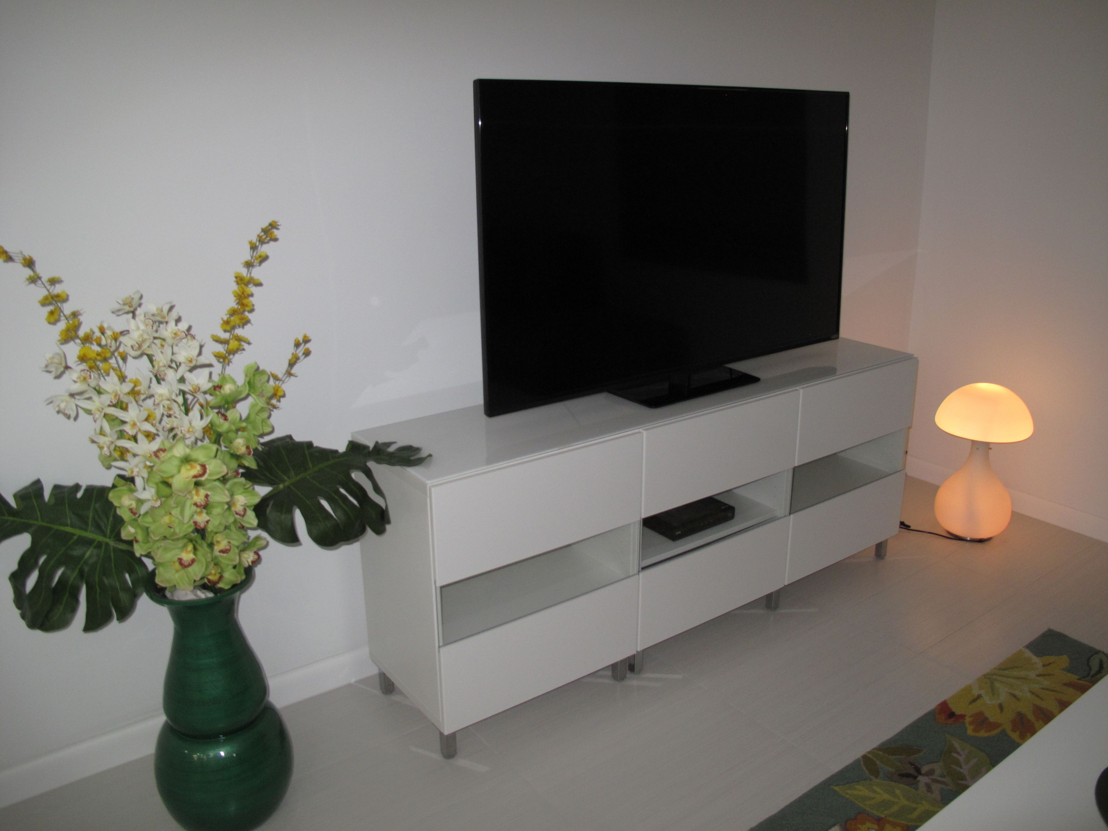 A modern living room with a flat-screen TV on a white cabinet, a green vase with flowers, and a floor lamp.