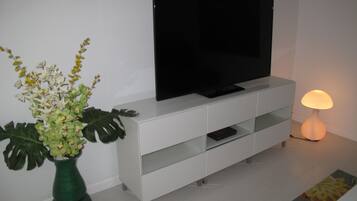 A modern living room with a flat-screen TV on a white cabinet, a green vase with flowers, and a floor lamp.
