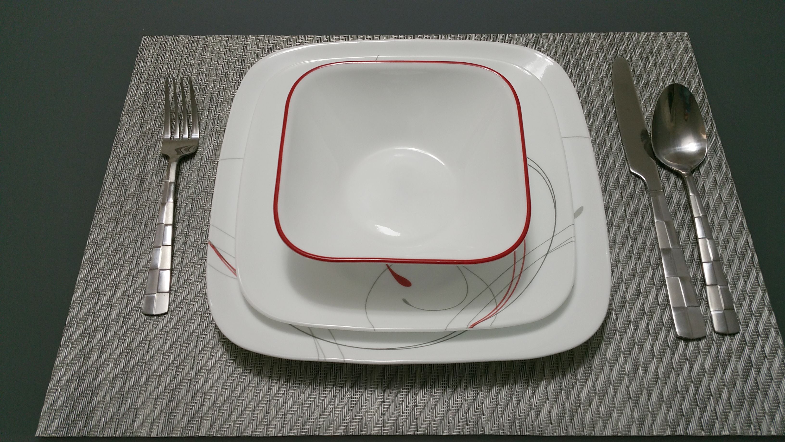 Une assiette carrée blanche avec un bord rouge, accompagnée d’une fourchette, d’un couteau et d’une cuillère, le tout posé sur un set de table texturé.