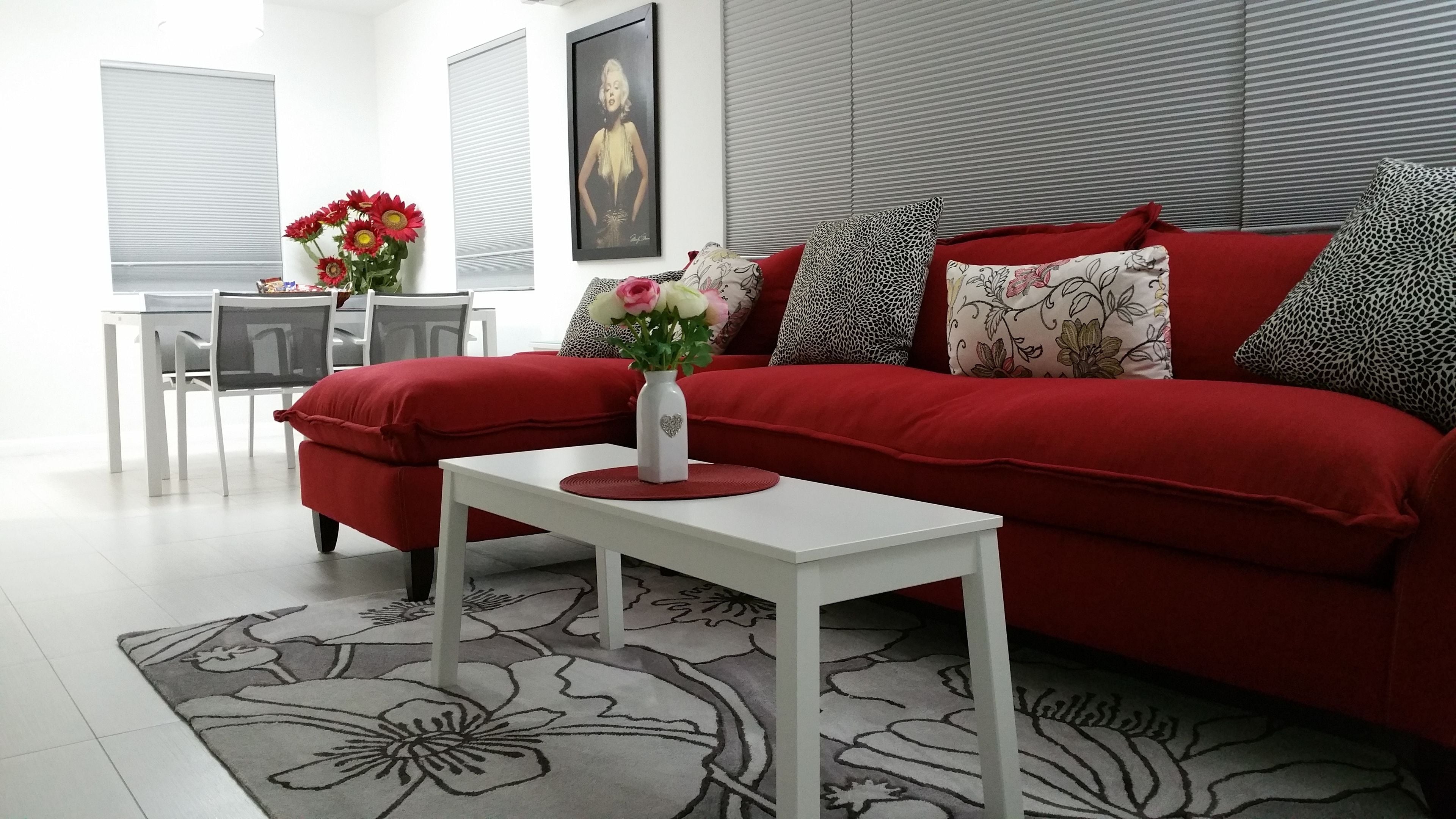 Un salon moderne avec un canapé rouge, une table basse blanche et un tapis à motifs floraux. On y trouve une petite table sur laquelle est posé un vase de fleurs, ainsi qu’un tableau encadré accroché au mur.
