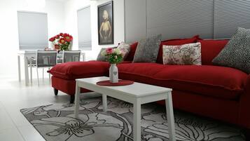 Un salon moderne avec un canapé rouge, une table basse blanche et un tapis à motifs floraux. On y trouve une petite table sur laquelle est posé un vase de fleurs, ainsi qu’un tableau encadré accroché au mur.