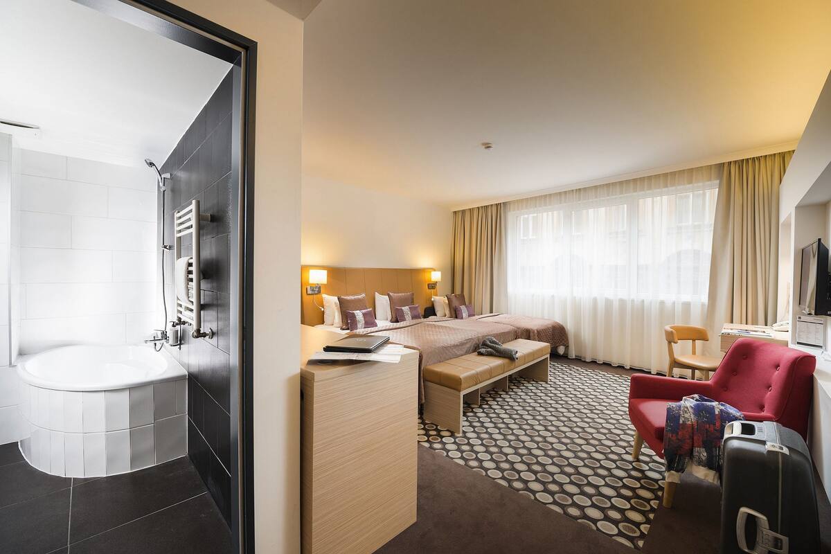 A hotel room with a bathtub, a bed with a bench, a red armchair, a desk, and a television.