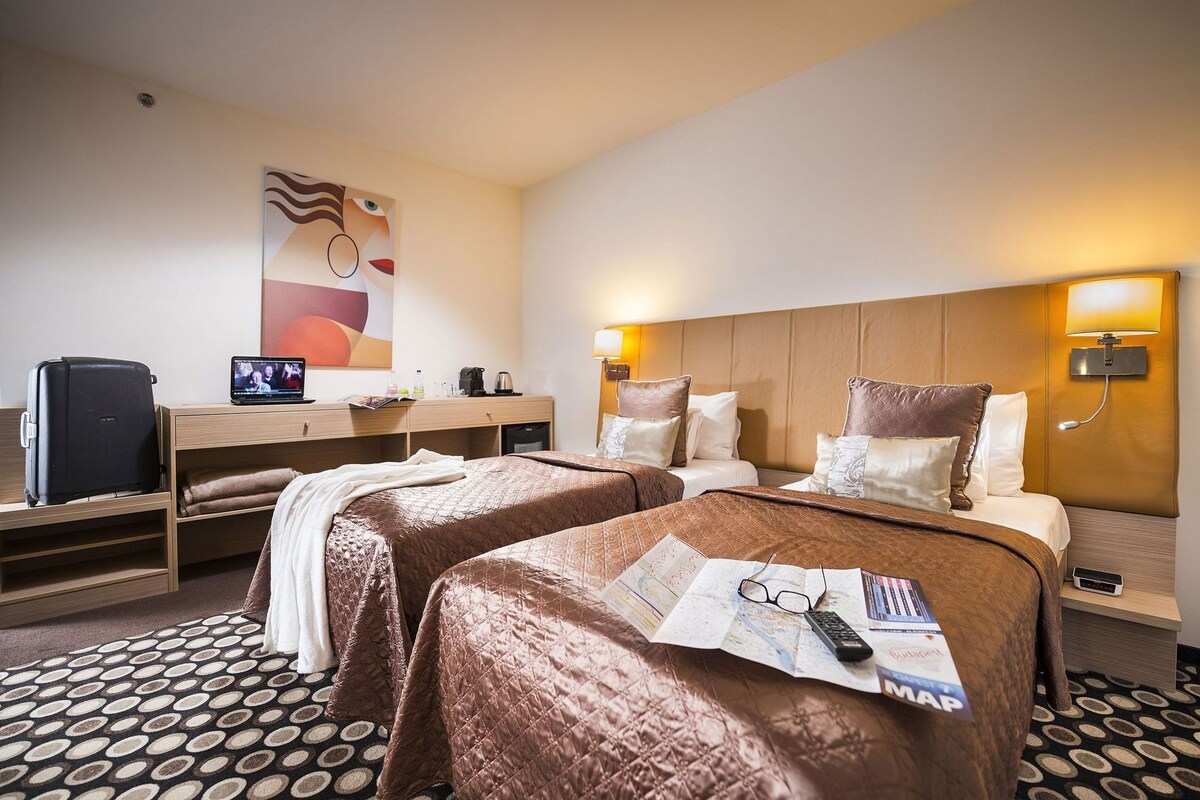 A hotel room with two beds, a desk with a laptop, a TV, and a wall-mounted lamp.