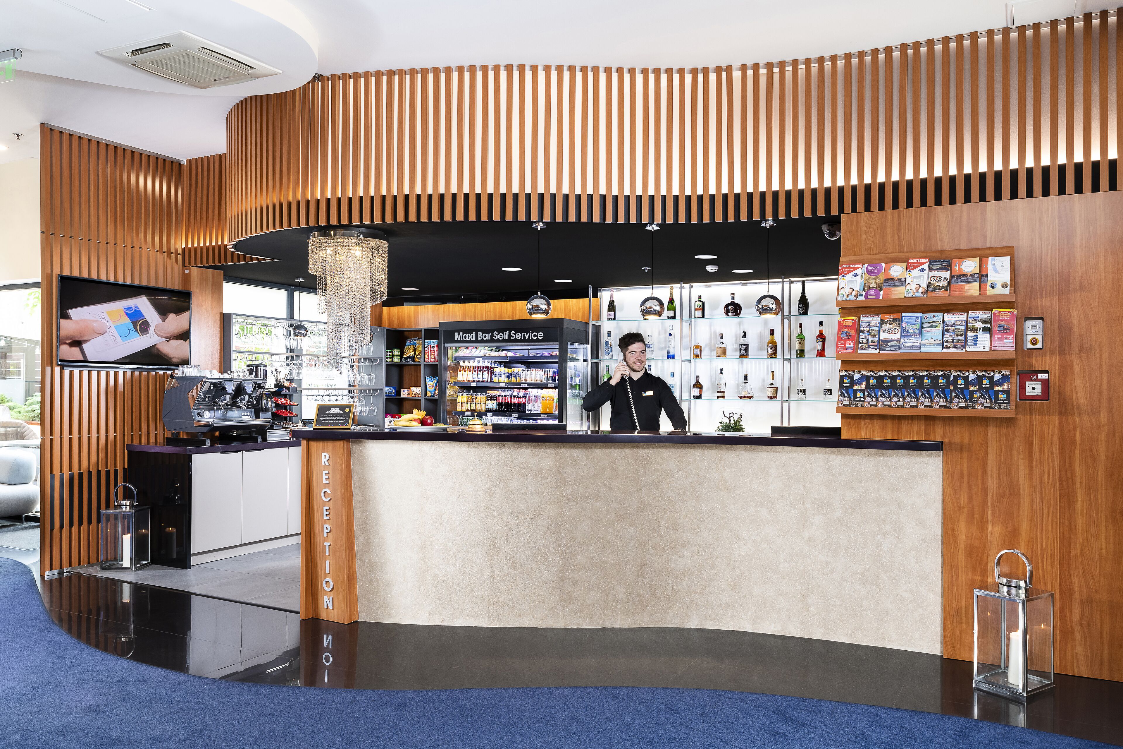 A modern reception area with a wooden reception desk, a self-service bar, and a display of magazines and books.