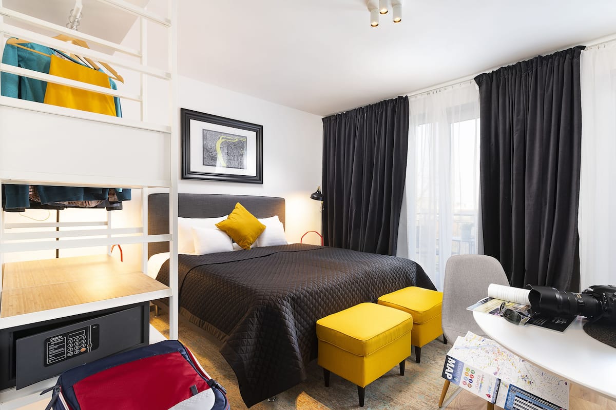 A hotel room with a bed, a desk with a camera, a chair, a desk lamp, a bookshelf, and a window with curtains.