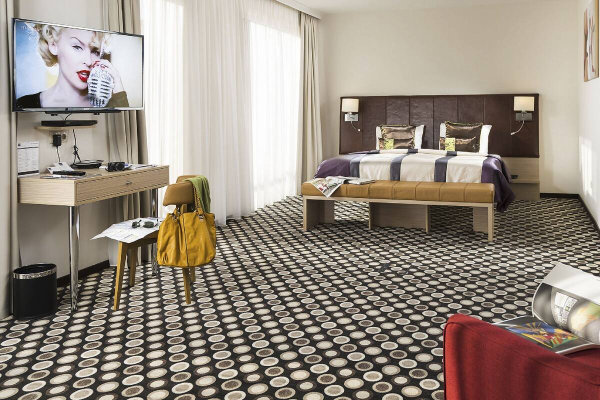 A hotel room with a bed, a desk, a television, a red armchair, and a patterned floor.