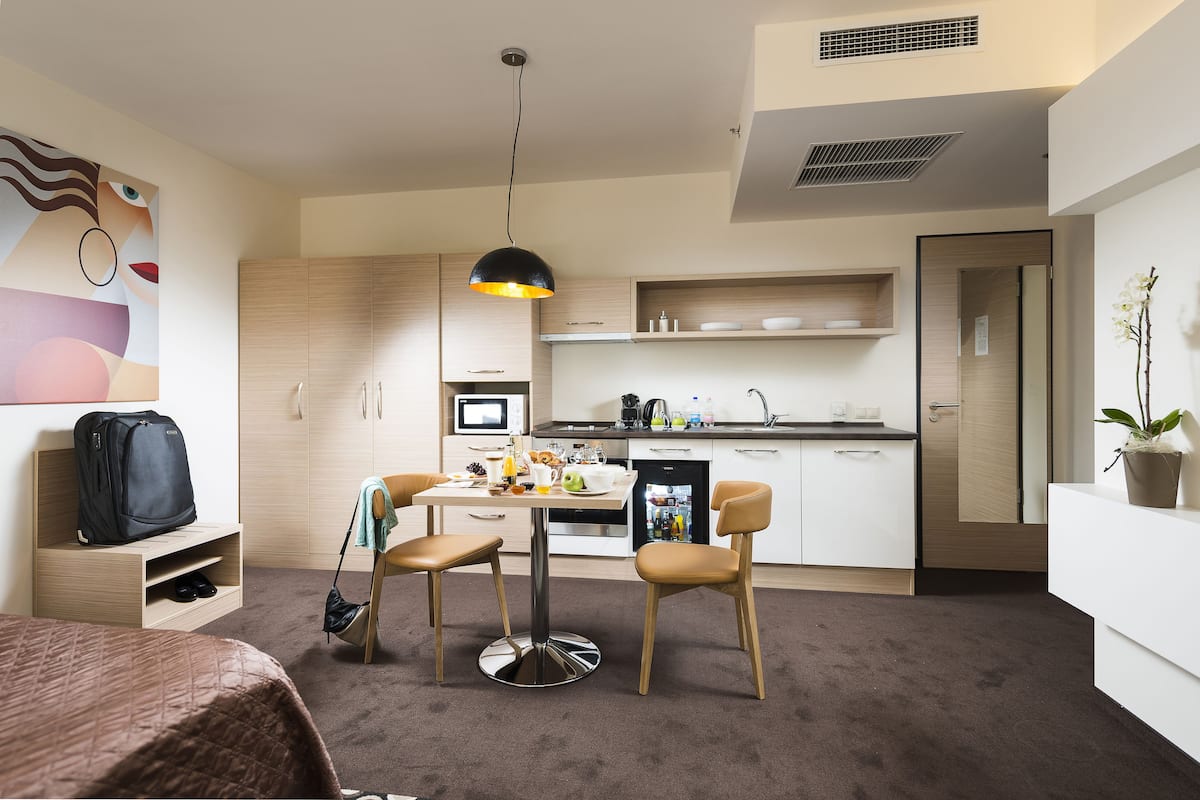 A compact hotel room with a kitchenette, a dining area, a bed, and a wall-mounted artwork.
