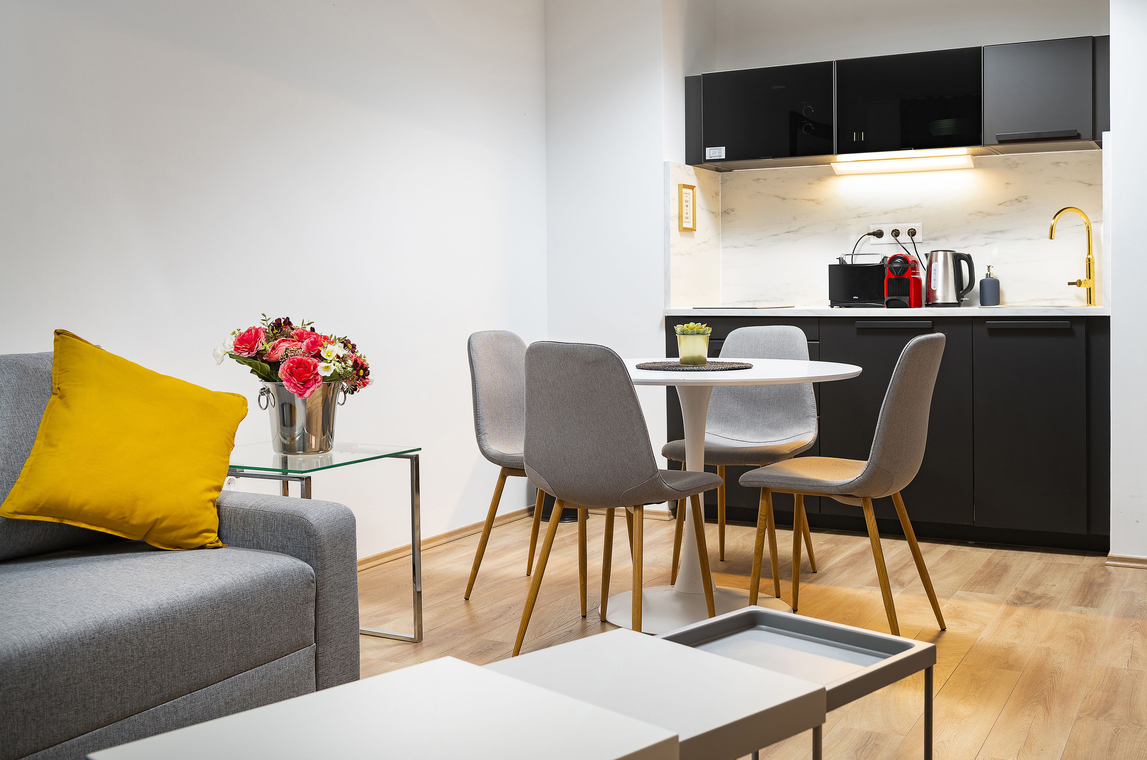 Ett modernt vardagsrum med en grå soffa, en gul kudde och en vas med blommor. En matplats med grå stolar och ett vitt bord syns. Köksdelen har svarta skåp, en vit bänkskiva och en guldgul kran.